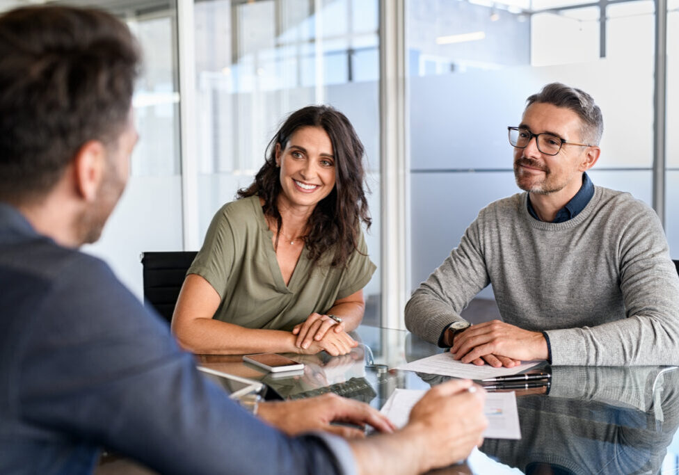 Smiling mature couple meeting with bank manager for investment. Beautiful mid adult woman with husband listening to businessman during meeting in conference room in modern office. Happy middle aged couple meeting loan advisor to buy a new home.